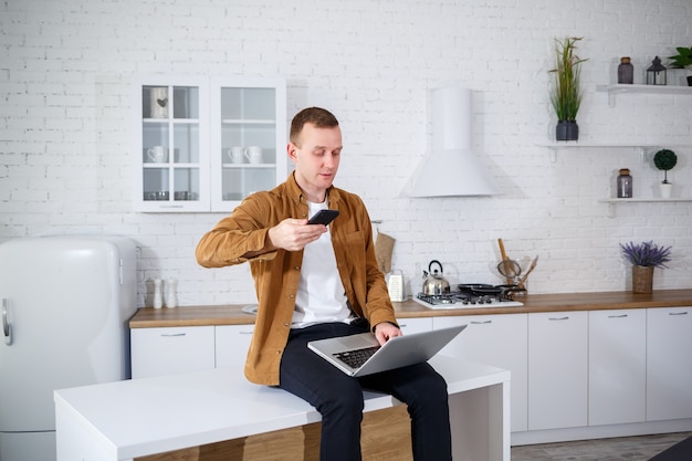 Um jovem atraente com roupas casuais, sentado na cozinha usando um computador laptop. Trabalhe em casa, fluxo de trabalho remoto.