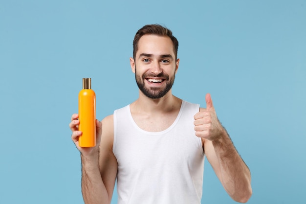 Um jovem barbudo de 20 anos de idade na camisa branca segura um frasco de xampu isolado no fundo da parede pastel azul, retrato de estúdio. Conceito de procedimentos cosméticos de saúde de cuidados com a pele. Simule o espaço da cópia.