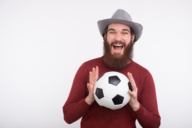 Um jovem barbudo feliz está segurando com as duas mãos uma bola de futebol perto de uma parede branca.