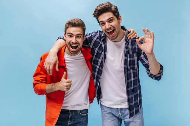 Foto um jovem barbudo legal de jaqueta laranja e moreno de camisa quadriculada sorrindo mostra polegar para cima e sinal de ok em fundo azul