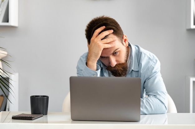 Um jovem barbudo pensativo e cansado sentado à mesa a trabalhar num portátil.