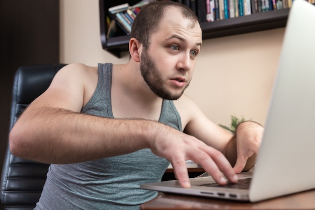 Um jovem barbudo vestindo uma camiseta cinza com roupas de casa usa laptop