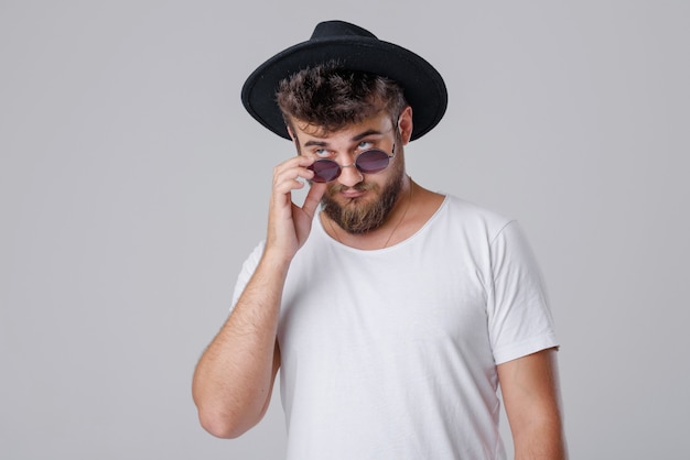 Um jovem bem sucedido com uma barba em óculos de sol e um chapéu em uma parede cinza