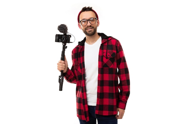 Um jovem blogueiro em um fundo branco em uma camisa xadrez e óculos para visão segura uma câmera com um