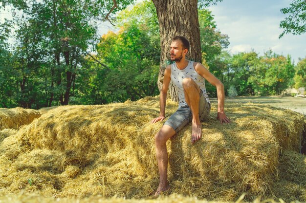 Um jovem bonito com barba de construção esbelta senta-se no feno e segura o feno nas mãos contra o fundo das árvores verdes no parque