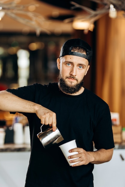 Um jovem bonito com barba, vestido em estilo casual, adiciona leite ao café em uma cafeteria moderna. .