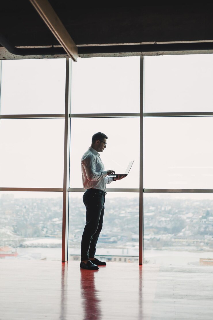 Um jovem bonito com um laptop está no fundo de uma grande janela panorâmica no céu Um escritório moderno com grandes janelas e um trabalhador de escritório
