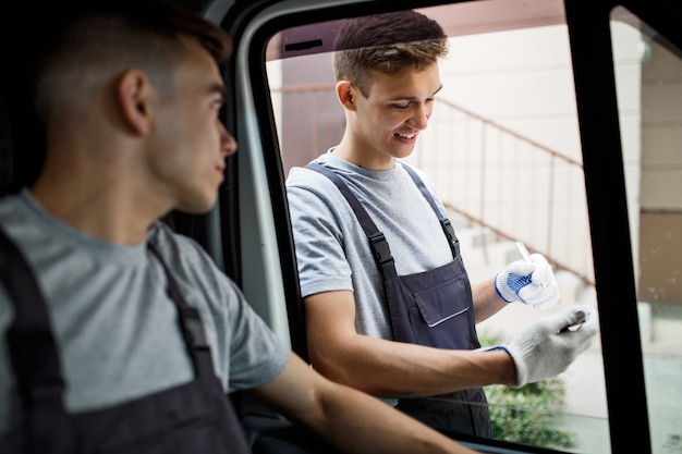 Um jovem bonito de uniforme está olhando pela janela do carro. Outro trabalhador de uniforme está olhando para a prancheta. Mudança de casa, serviço de mudança.