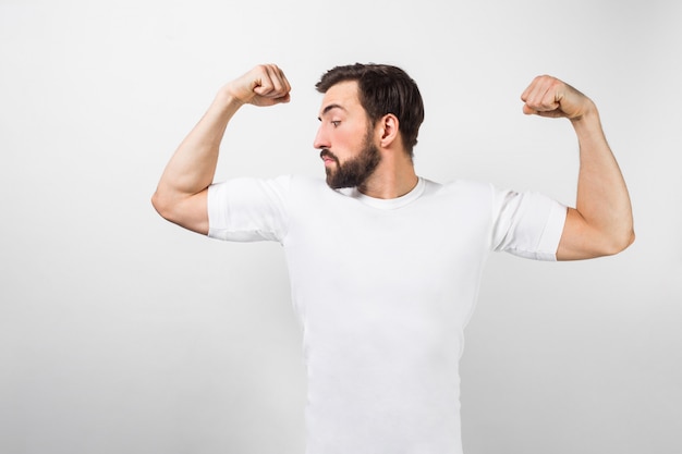 Um jovem bonito e confiante em pé e mostrando grandes músculos nas mãos. ele está olhando para um deles e muito orgulhoso deles. ele está vestindo uma camiseta branca, isolada na parede branca.