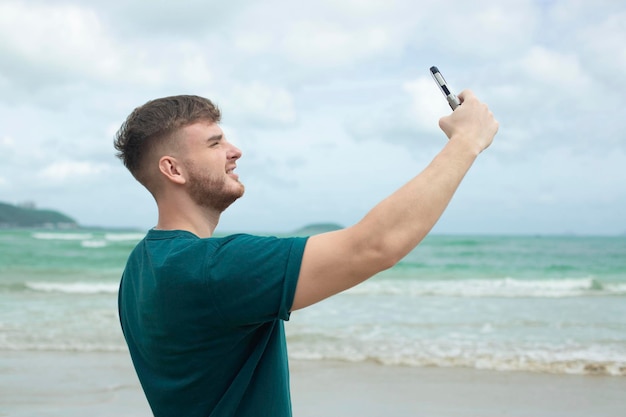 Um jovem bonito e feliz no mar está tirando uma foto de si mesmo selfie na câmera de seu telefone usando smartphone para mídia social na praia de verão em um país exótico tropical Blogger vlog