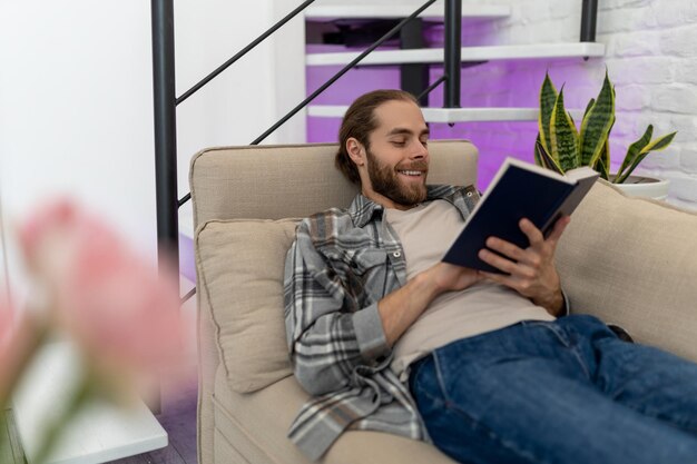 Um jovem bonito e sorridente a ler um livro sentado no sofá da sala de estar.