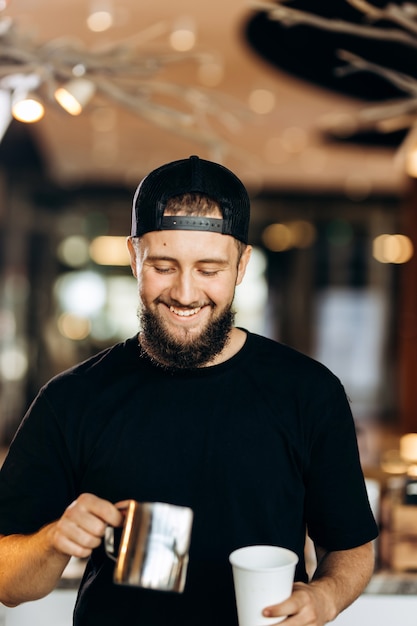 Um jovem bonito e sorridente com barba, vestido em estilo casual, adiciona leite ao café em uma cafeteria moderna. .