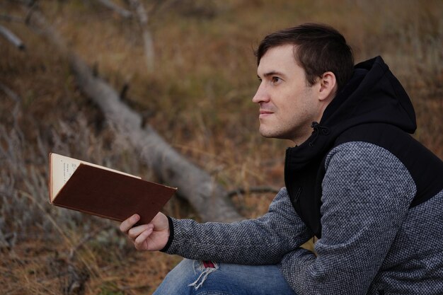 Foto um jovem bonito em roupas casuais está sentado em um tronco e lendo um livro na floresta de outono