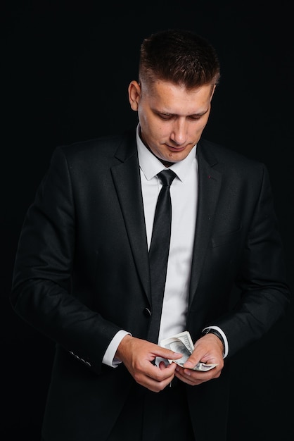 Um jovem bonito em um elegante terno preto joga dólares em um fundo preto. Um homem de negócios elegante. Um grande empresário.