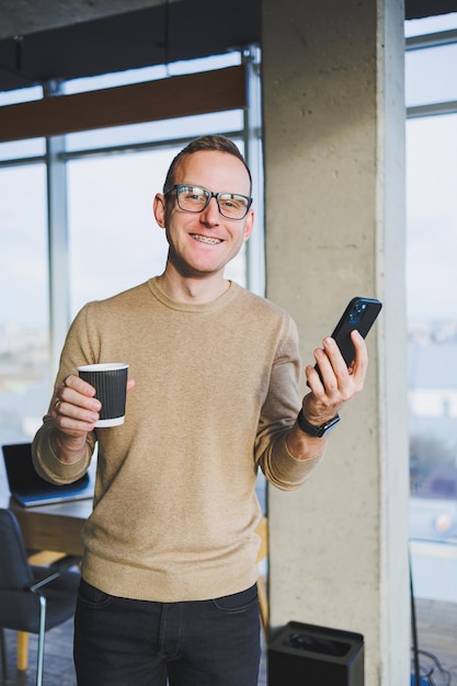 Um jovem bonito em um suéter marrom e óculos está falando emocionalmente em um telefone celular e bebendo café enquanto relaxa no escritório Um jovem freelancer trabalha remotamente