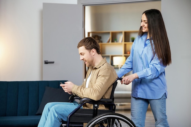 Um jovem bonito em uma cadeira de rodas está segurando um telefone celular enquanto sua namorada o ajuda a se mover pelo apartamento