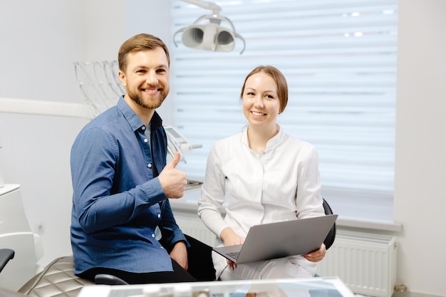 Um jovem bonito está conversando com uma médica em uma consulta odontológica em um belo escritório brilhante O dentista explica ao paciente e mostra tudo no laptop