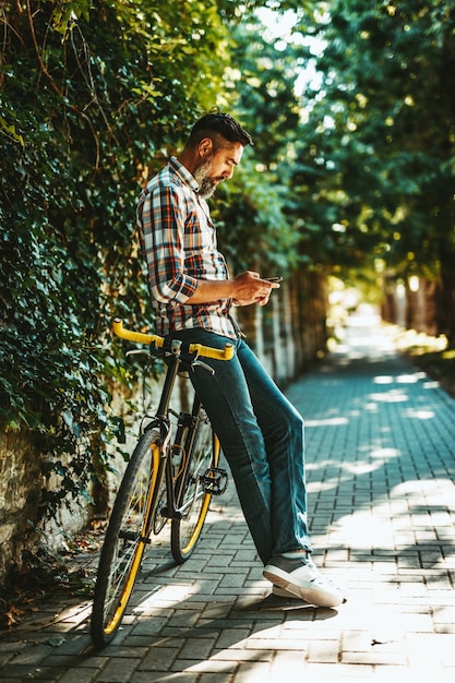 Um jovem bonito vai para a cidade com sua bicicleta, parado ao lado dela, esperando alguém e enviando mensagem de texto.