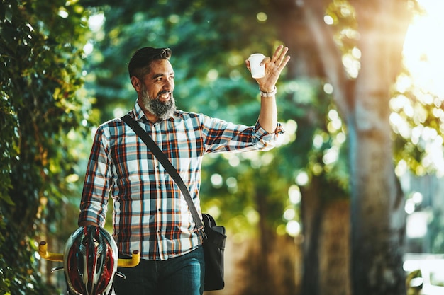 Um jovem bonito vai para o passeio da cidade com sua bicicleta, de pé ao lado dela e acenando para alguém.