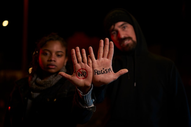 Um jovem branco e uma jovem negra exibindo uma mensagem escrita à mão contra o racismo.