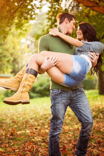 Um jovem carrega em seus braços uma jovem atraente em um parque, andando sobre folhas caídas.