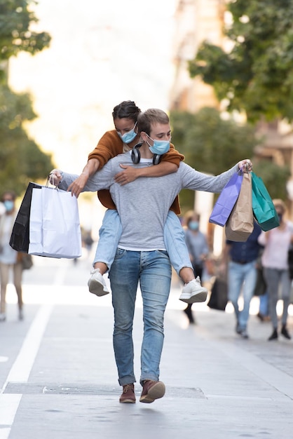 Um jovem carregando sua namorada nas costas com algumas sacolas de compras usando máscaras médicas