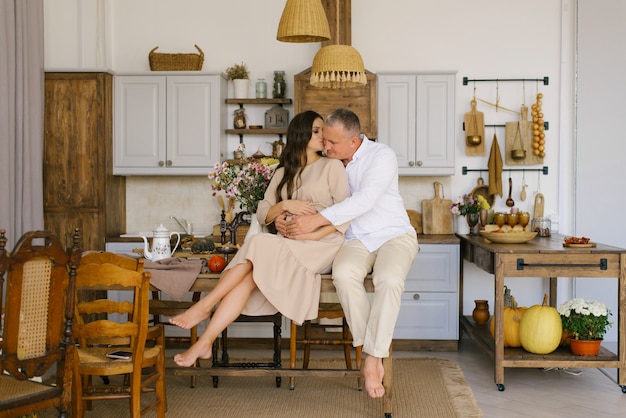 Um jovem casal abraços enquanto está sentado em uma mesa na cozinha. O marido abraça a esposa grávida, colocando as mãos na barriga grande dela. O estilo de vida da família