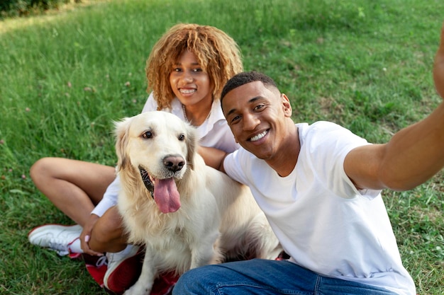 Um jovem casal afro-americano senta-se no parque com o cão e tira uma selfie.