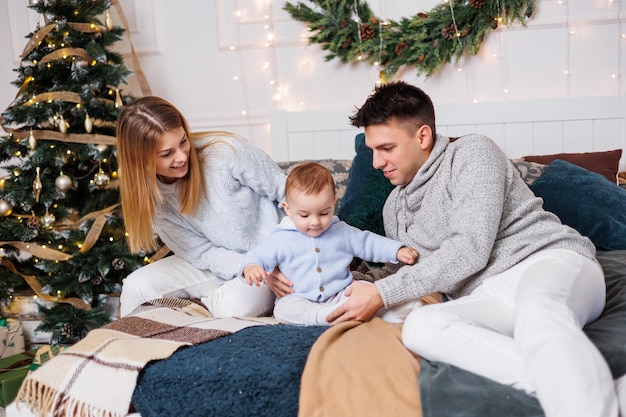 Um jovem casal alegre com um filho pequeno está brincando na cama perto da árvore de Natal Interior de Ano Novo no quarto Árvore de Natal com brinquedos Atmosfera familiar festiva