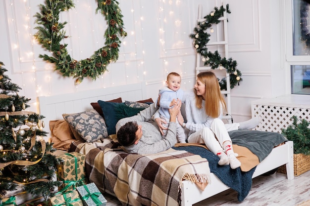 Um jovem casal alegre com um filho pequeno está brincando na cama perto da árvore de Natal Interior de Ano Novo no quarto Árvore de Natal com brinquedos Atmosfera familiar festiva