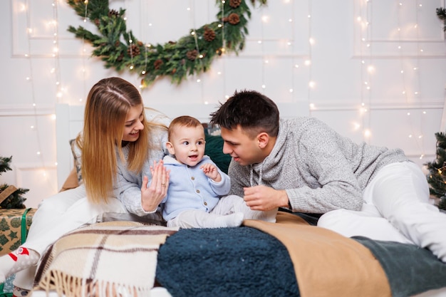 Um jovem casal alegre com um filho pequeno está brincando na cama perto da árvore de Natal Interior de Ano Novo no quarto Árvore de Natal com brinquedos Atmosfera familiar festiva