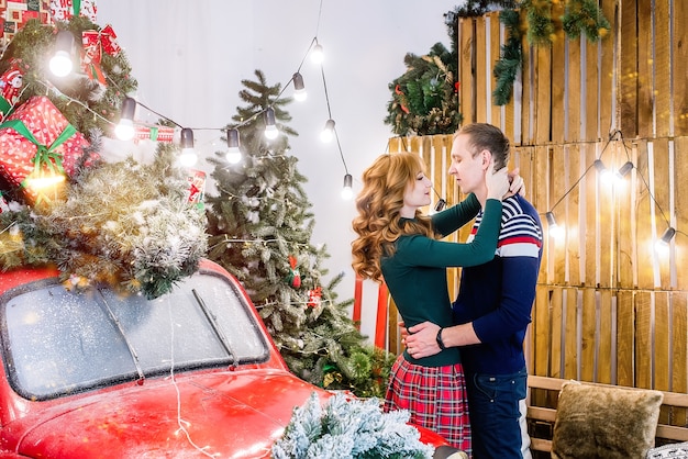 um jovem casal apaixonado contra árvores de Natal com guirlandas. Um jovem abraça uma mulher perto do carro vermelho.