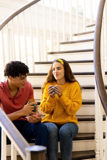 Um jovem casal biracial segurando copos de café e conversando enquanto está sentado na escada em casa, espaço de cópia