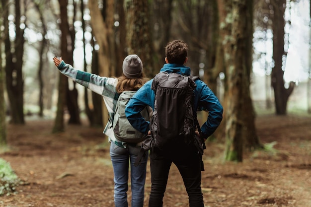 Um jovem casal caucasiano de casacos caminha na floresta juntos apontando o dedo para o espaço vazio. Aproveite a viagem.