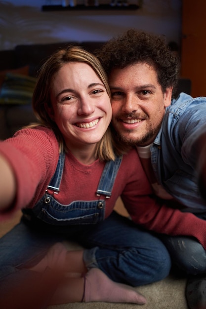 Um jovem casal caucasiano feliz vertical fazendo uma selfie sorridente olhando para a câmera em casa à noite