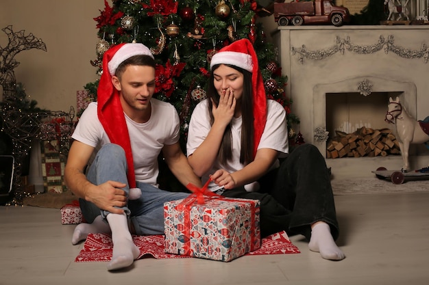 Foto um jovem casal com chapéu de papai noel está sentado no chão perto da árvore de natal ao lado de caixas de presente