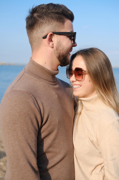 Um jovem casal de suéteres jeans e óculos escuros fica na praia à beira do lago e dá as mãos