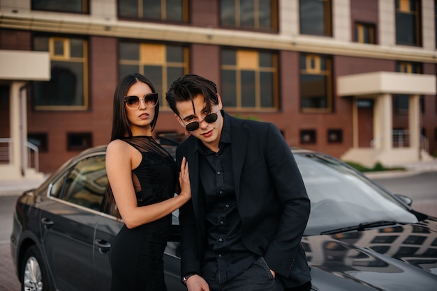 Foto um jovem casal elegante de preto fica perto do carro ao pôr do sol. moda e estilo