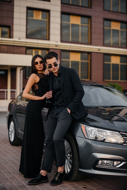 Um jovem casal elegante de preto fica perto do carro ao pôr do sol. Moda e estilo.