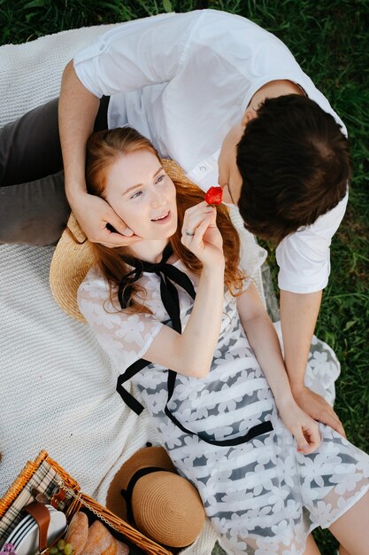 Um jovem casal está descansando no parque em um cobertor comendo morangos frescos, uma garota romântica e um cara fazem um piquenique ao ar livre fora da cidade