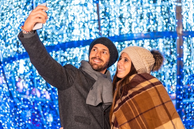 Um jovem casal europeu curtindo as luzes de Natal da cidade tirando uma selfie com o celular para as redes sociais