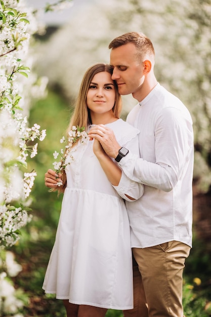 Um jovem casal feliz apaixonado fica em um jardim de macieiras florescendo. Um homem de camisa branca e uma garota de vestido branco claro estão andando em um parque florido