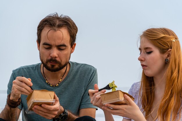 Foto um jovem casal focado a comer comida para levar enquanto está sentado na rua.