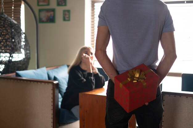 Um jovem casal, homem segurando uma caixa de presente nas costas, fazendo surpresa para a namorada