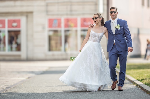 Um jovem casal recém-casado andando feliz na cidade em vestes de casamento com um lindo buquê.