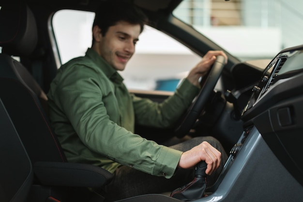 Um jovem caucasiano alegre testa um carro novo antes da compra no foco seletivo da concessionária de automóveis