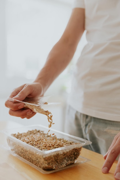 Um jovem caucasiano irreconhecível derrama sementes de um saco transparente em um recipiente com terra na cozinha, em uma mesa de madeira, vista lateral