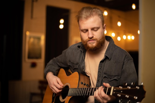 Um jovem com barba toca violão em uma sala com iluminação quente