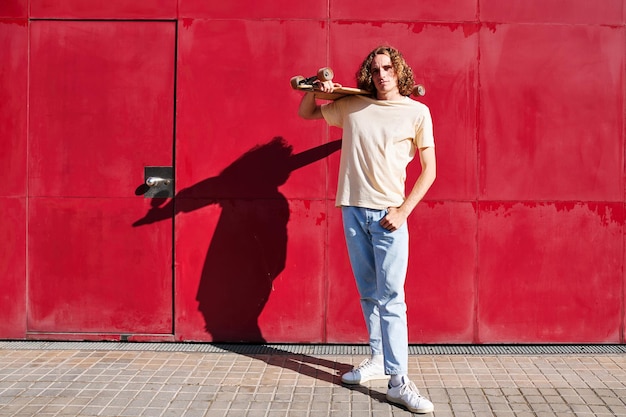 Um jovem com cabelo encaracolado e seu skate com um fundo vermelho em um dia ensolarado de verão