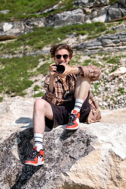 Um jovem com cabelo encaracolado tirando fotos com a câmera na natureza, sentado na encosta de uma colina rochosa